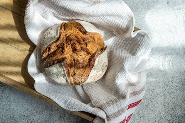 A freshly baked sourdough loaf made with rye flour, resting on a wooden board covered with a white kitchen towel Model Released AnnaBogush_ID21101_622339_004 Copyright: xAnnaxBogushx