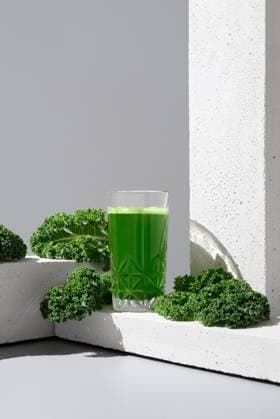A transparent glass filled with vibrant green kale juice is placed next to fresh, crisp kale leaves on a modern white surface, showcasing a healthful drink option Model Released RuthCarrodeguas_ID21186_624725_005 Copyright: xRuthxCarrodeguasx RECORD DATE NOT STATED
