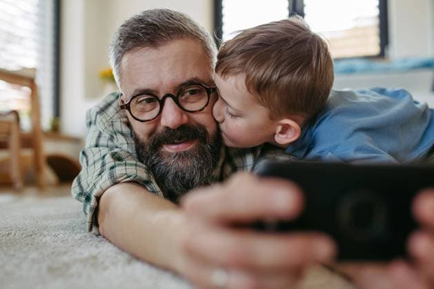 Little boy making selfie on smartphone with father, lying on floor in kids room, making silly faces. Dad explaining technology to son, digital literacy for kids. Little boy making selfie on smartphone with father, lying on the floor in kids room, making silly faces. Dad explaining technology to son, digital literacy for kids. model released