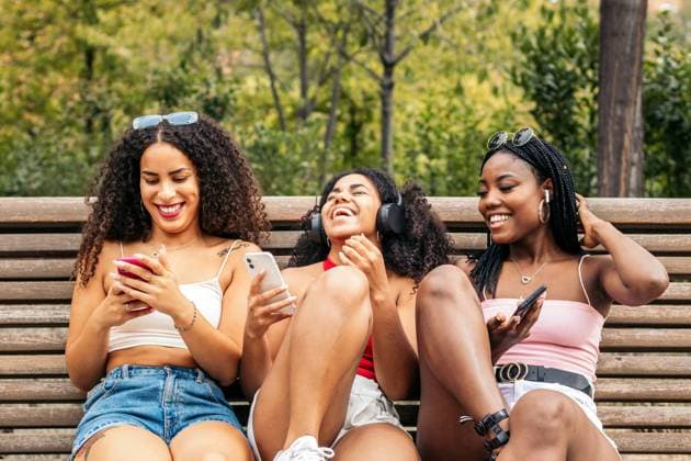 IMAGO Creative: Sommer, Sommertage im Stadtpark African american female friends sitting on a park bench laughing and having fun while using smartphone and listening to music with headphones. Model Released PedroMerino_ID18922_563471_005 Copyright: xPEDROxMERINOx