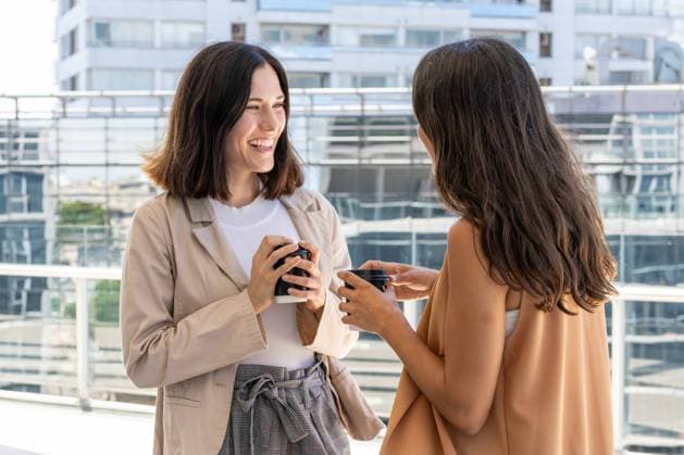 Two businesswomen are standing outside a building, enjoying their office break. They are smiling and holding cups of coffee, indicating a sense of camaraderie and friendship. Model Released ValeriaVenezia_ID20934_617621_025 Copyright: xx