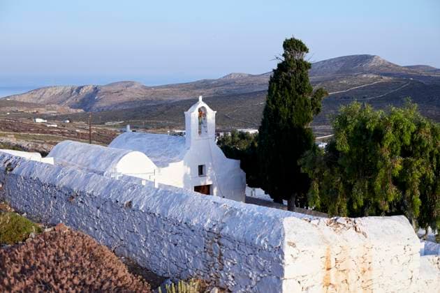 A serene landscape featuring a traditional white-washed church with a bell tower, surrounded by a stone wall, overlooking a hilly terrain with the sea in the distance at dawn. Folegandros Greece bola01005 Copyright: xConnectxImagesx