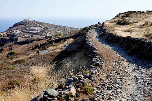 A stone hiking trail leads through a dry, grassy landscape toward a distant overlook with a clear blue sky above. Folegandros Greece bola01011 Copyright: xConnectxImagesx