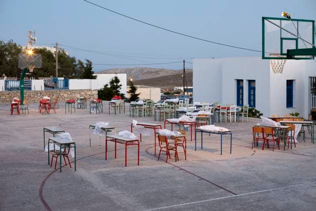 An outdoor setting at dusk featuring a basketball court with tables and chairs neatly arranged across it, under the glow of gentle lights, adjacent to a white building with blue trimmings. Folegandros Greece bola01016 Copyright: xConnectxImagesx