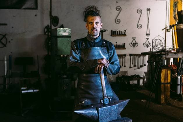 Creative Highlights Symbolbilder A male blacksmith with a contemplative expression stands looking ay camera in a dimly lit smithy, leaning on his anvil with hammers nearby. Model Released DavidBenito_ID21451_631182_075 Copyright: xDavidxBenitox