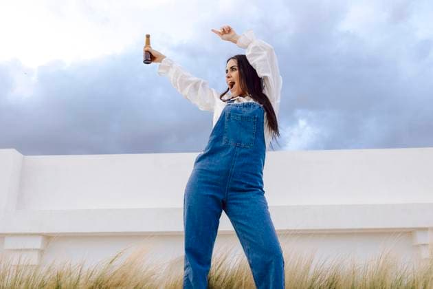Creative Highlights Symbolbilder From below joyful woman in denim overalls holding a beer bottle aloft in a festive pose at the port, with cloudy sky overhead. Model Released LuisTenza_ID21766_638614_027 Copyright: xLuisxTenzax RECORD DATE NOT STATED