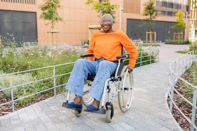 IMAGO Creative: Sommer, Sommertage im Stadtpark Black woman with a bright smile, dressed in orange top and jeans, navigates a wheelchair through a serene park, exemplifying accessibility and joy Model Released DavidMunoz_ID22272_651064_052 Copyright: xDavidxMunozx