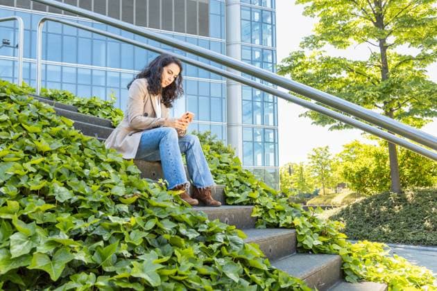 IMAGO Creative, Lifestyle, Frühling in der Stadt A professional woman in casual business attire is seated on steps overgrown with lush greenery, focused on her smartphone, with a modern glass office building in the background Model Released IgnacioFerrandiz_ID22117_647152_027 Copyright: xIgnacioxFerrandizx RECORD DATE NOT STATED