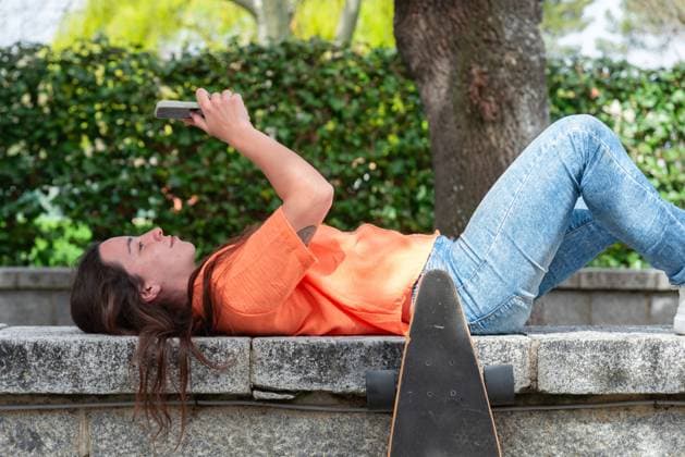 IMAGO Creative, Lifestyle, Frühling in der Stadt A young woman in a casual orange shirt and jeans lies on a stone bench with her skateboard propped beside her. She is comfortably looking at her smartphone, surrounded by lush, green foliage in a park setting Model Released NataliaPenado_ID20782_613182_013 Copyright: xNataliaxPenadox RECORD DATE NOT STATED