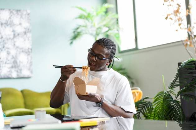 A black man sits in a comfortable indoor setting, using chopsticks to eat noodles from a takeout box Model Released VictorCuenca_ID22085_646425_028 Copyright: xVictorxCuencax