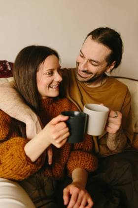 A Caucasian woman and man sitting closely on a cream-colored sofa, sharing a tender gaze. The woman, holding the man s hand, lovingly looks at him while he, with a subtle smile. They each hold a cup, adding a relaxed, homely vibe. Model Released CamilleGirard_ID21717_637085_023 Copyright: xCamillexGirardx