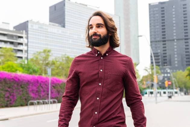 A stylish young man with a beard walks confidently on a tree-lined street in Poblenou, Barcelona, dressed in a smart burgundy shirt, appreciating the modern architecture and vibrant city life around him Model Released DavidMunoz_ID22411_654778_004 Copyright: xDavidxMunozx