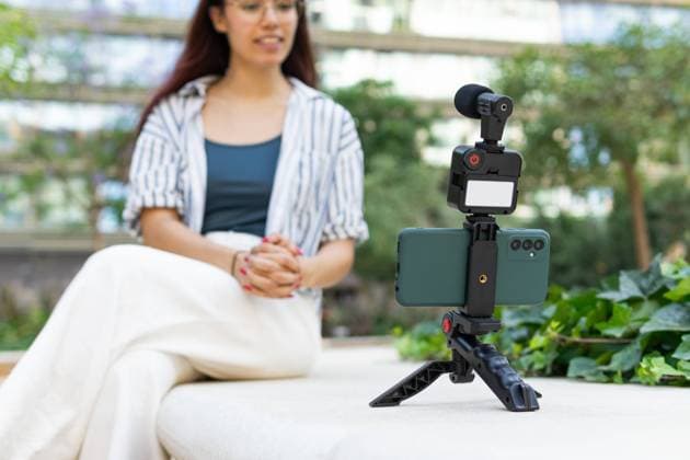A young woman with redhead, wearing a striped shirt and white pants, is recording herself using a smartphone mounted on a tripod, with a blurred green park in the background Model Released DavidMunoz_ID22797_663737_026 Copyright: xDavidxMunozx