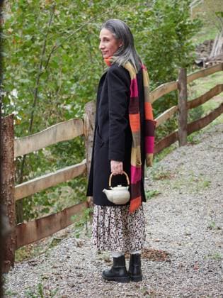 A mature woman with gray hair, smiling gently, holds a tea pot during an autumn walk. She wears a stylish black coat and a colorful scarf, gazing away from the camera Model Released AnaLopez_ID22966_667622_002 Copyright: xAnaxLopezx RECORD DATE NOT STATED