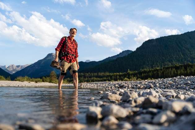 Senior man hiking through a rocky stream in the mountains with a backpack, flannel shirt, and shorts on a sunny day. model released, Symbolfoto, DIGF21510
