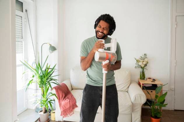 IMAGO Creative, Frühling, Frühjahrsputz Happy man singing with vacuum cleaner standing in living room at home model released, Symbolfoto property released, VRAF00688
