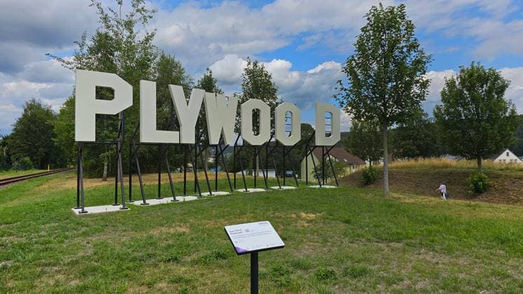 Skulptur_Plywood 23.07.2024, Olbernhau, Erzgebirge, 38 Kommunen und Gemeinden bilden gemeinsam mit Chemnitz die Kulturhauptstadt-Region, die im Jahr 2025 Europäische Kulturhauptstadt sein wird. Bis zum Jahr 2025 und darüber hinaus wird der PURPLE PATH, ein großer Skulpturen-, Kunst- und Macherweg und Flagship der Kulturhauptstadt, in der Region entstehen. Parkplatz am Bahnübergang Rübenauer Weg Brandauer Straße in Höhe Bahnhof Olbernhau, Die in weißem Farbton pulverbeschichtete, aus Stahlblechen gefertigte Skulptur des 1984 geborenen, in Karl-Marx-Stadt Chemnitz aufgewachsenen und heute in Berlin lebenden Künstlers Jay Gard lässt das Wort Plywood (Sperrholz) lesen. Metall simuliert Holz, verstärkt wird das Verwirrspiel