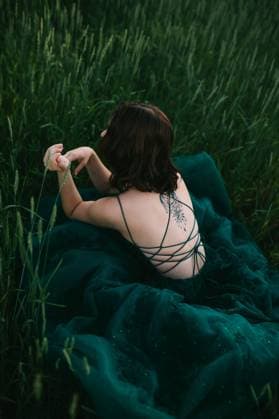 Creative Highlights Symbolbilder A serene image capturing a young woman sitting in a field, wearing an elegant dark green prom dress with intricate back straps and a tattoo visible on her shoulder Model Released CamilleGirard_ID13413_408647_026 Copyright: xCamillexGirardx