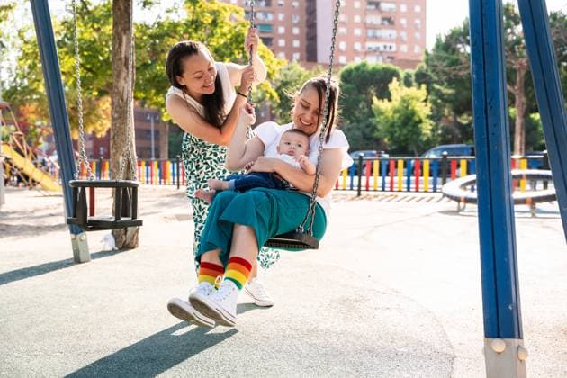 IMAGO Creative, Diversity, Familie: Gleichgeschlechtliche Eltern: Zwei Mütter A joyful lesbian couple smiles and engages with their baby son while playing on a park swing. One mother gently pushes the swing, while the other holds their child, capturing a moment of family love and happiness outdoors. Model Released ManuelRuiz_ID23737_688702_022 Copyright: xManuelxRuizx