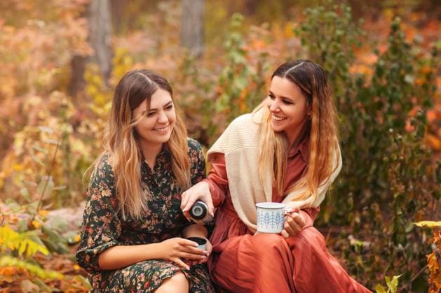 Junge Freundinnen in stilvollen Kleidern schauen sich an und genießen ein heißes Getränk, während sie auf Kürbissen im Herbstwald sitzen Young female friends in stylish dresses looking at each other and enjoying hot beverage while sitting on pumpkins in autumn forest Copyright: imageBROKER DashaxPetrenko ibxiqx09193662.jpg ,model released, Symbolfoto ,property released