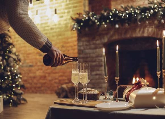 Silvesterfeier. Mann gießt Champagner in Gläser stehen auf dem Tisch mit festlichen Weihnachtsessen, Kerzen und eingepackt Geschenke gegen unscharfen Hintergrund von Backstein Kamin und firtree, beschnitten New Years Eve celebration. Man pouring champagne into glasses standing on table with festive xmas dinner, candles and wrapped gifts against blurred background of brick fireplace and firtree, cropped Copyright: imageBROKER DashaxPetrenko ibxiqx09201485.jpg