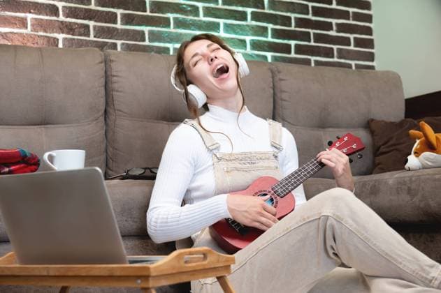 Musikunterricht aus der Ferne, Ukulele spielen oder Gesangsunterricht, Singen. Hipster Schüler, der eine Unterrichtsstunde aufnimmt oder mit einer Laptop Webcam lernt, während er auf dem Boden im Wohnzimmer sitzt Remote music lessons, playing the ukulele or teaching vocals, singing. Hipster student recording lesson or studying using laptop webcam while sitting on floor in living room Copyright: imageBROKER IanxIankovskii ibxiqx09619627.jpg ,model released, Symbolfoto ,property released