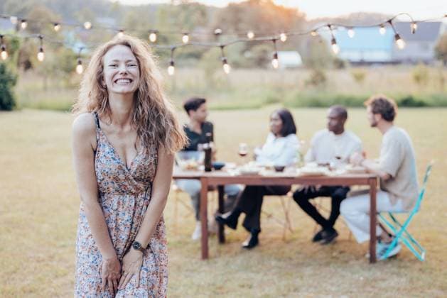 Creative Highlights Symbolfotos Joyful Woman at Outdoor Garden Party This image features a young woman in the foreground, standing in a grassy outdoor area adorned with string lights above. She is wearing a floral dress and is smiling broadly, showcasing a look of genuine happiness and laughter. Her curly hair is tousled, adding to her carefree and joyful appearance. In the background, a diverse group of people can be seen seated around a table, engaged in conversation, suggesting a social gathering or celebration among friends. Joyful Woman at Outdoor Garden Party. High quality photo ,model released, Symbolfoto Copyright: xZoonar.com BjornxBeheydtx 21299502 ,model released, Symbolfoto ,property released