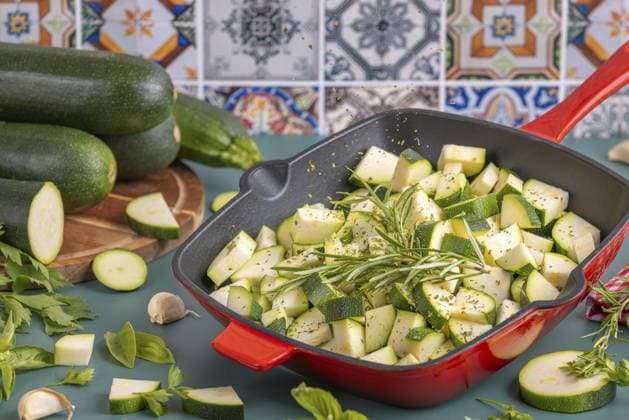 Diced courgettes in a red pan, flying herbs, stacked courgettes in the background in front of colourful tiles Copyright: imageBROKER LucasxSeebacher ibxlse11007864.jpg