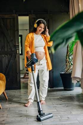 IMAGO Creative, Frühling, Frühjahrsputz Happy pregnant woman wearing headphones and using vacuum cleaner at home model released, Symbolfoto property released, VSNF02373