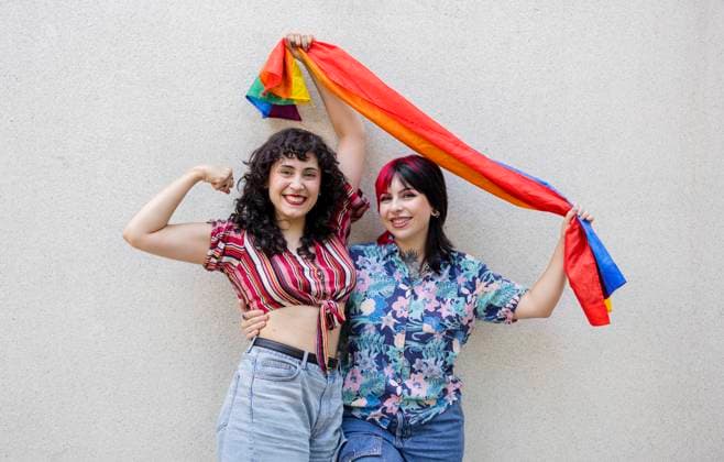 IMAGO Creative, Diversity, Teenager: LGBTQIA+ Creative Highlights Symbolfotos Two joyful lesbian friends expressing happiness and pride while holding a colorful rainbow flag against a neutral background, symbolizing LGBTQIA+ unity and celebration Model Released JoseCarlosIchiro_ID23746_689118_029 Copyright: xJosexCarlosxIchirox