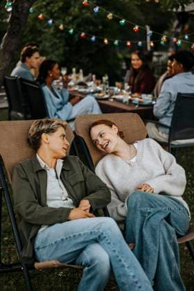 IMAGO Creative, Lifestyle, Feste im Garten #2 Female friends with legs crossed at knee sitting on chair and talking in back yard at social party Sweden, Stockholm, model released, property released Copyright: xMaskotx, MA112455