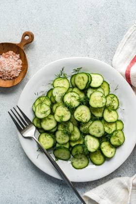 Top view of a fresh cucumber salad topped with dill, served in a white bowl alongside a wooden spoon of pink sea salt and a grey textile napkin. Ideal for a healthy summer dish. Model Released AnnaBogush_ID24540_711383_006 Copyright: xAnnaxBogushx RECORD DATE NOT STATED