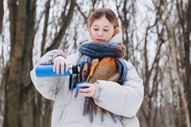 A young girl looks down while pouring a hot drink from a blue thermos into a cup, wearing a white winter coat and colorful scarves in a snowy forest setting. Model Released HannaYakimenko_ID24348_706112_008 Copyright: xHannaxYakimenkox