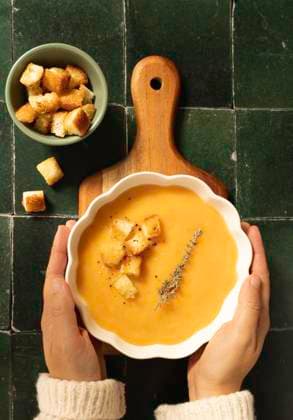 A top-view image of hands holding a stylish white bowl of creamy pumpkin soup garnished with croutons, fresh thyme, and a dash of black pepper, served on a wooden board over a green tiled surface Model Released MaiderAlvarez_ID24343_705989_006 Copyright: xMaiderxAlvarezx