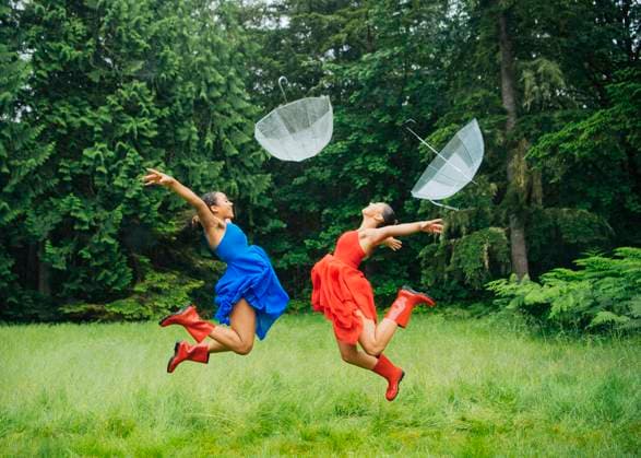 Creative Highlights Symbolfotos Two women in colorful dresses joyfully jumping with transparent umbrellas in a lush green forest clearing. sape00130 Copyright: xConnectxImagesx ,model released
