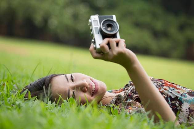 Smiling woman photographing through old camera lying down on grass at park model released, Symbolfoto, EAF00293