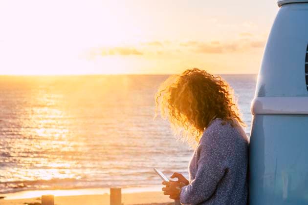 Woman using smart phone by beach at sunset in Fuerteventura, Canary Islands , 04.07.2019, model released, TTBB_ti0564_00080.jpg, Color Image,Horizontal,Focus On Foreground,Selective Focus,Outdoors,Sunset,Dusk,Fuerteventura,Canary Islands,Spain,Travel Destinations,Travel,One Person,People,40-44 Years,Mid Adult,One Mid Adult Woman Only,Only Women,Adult,Mid Adult Women,Caucasian Appearance,Personal Land Vehicle,Camper Van,Road Trip,Transportation,Getting Away From It All,Tourist,Beach,Sand,Coastline,Sea,Water,Blond Hair,Curly Hair,Sweater,Holding,Smart Phone,Communication,Sunbeam,Sunlight,Text Messaging,Mobile Phone,Wireless Technology,Cloud,Leaning Copyright: xFabioxCamandonax