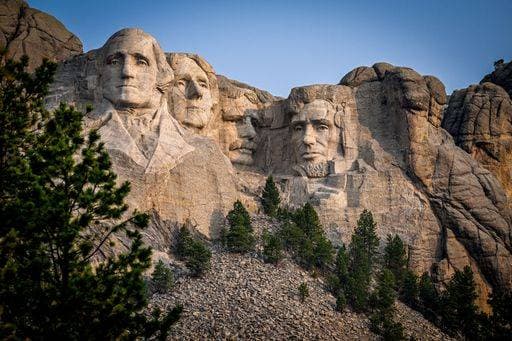 Mount Rushmore National Memorial with clear blue sky and surrounding pine trees. 25370795
