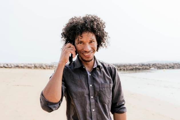 Creative Highlights Symbolfotos A joyful man with curly hair chats on his smartphone while standing on a sunny beach, wearing a dark casual shirt, embodying a carefree, adventurous vibe Model Released PaulaBerezo_ID24305_704389_011 Copyright: xPaulaxBerezox