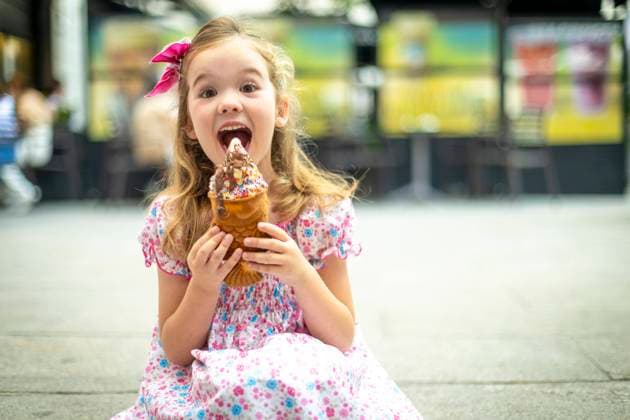 Creative Highlights Symbolfotos Joyful young girl with a pink bow and floral dress delightfully eating a large, colorful ice cream cone in an outdoor setting. Model Released SloomStudio_ID23813_691335_005 Copyright: xx RECORD DATE NOT STATED