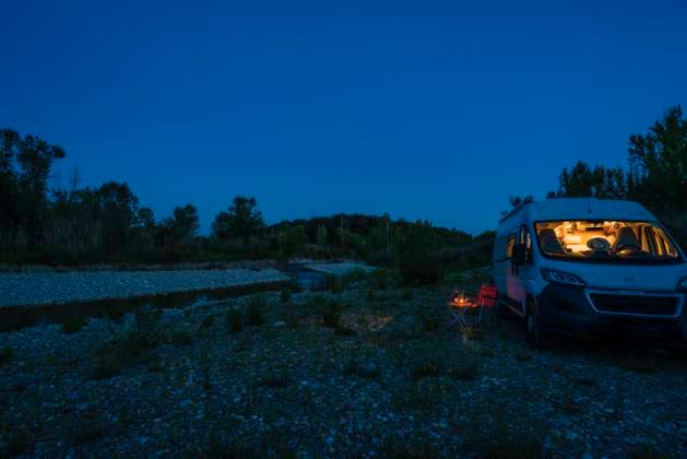 EIn Mann sitzt auf einem Campingstuhl vor einem Tisch an einem Sommerabend zur blauen Stunde vor seinem Camper. Deutschland Mit einem Campervan unterwegs *** A man sits on a camping chair in front of a table on a blue hour summer evening in front of his campervan Germany On the road in a campervan