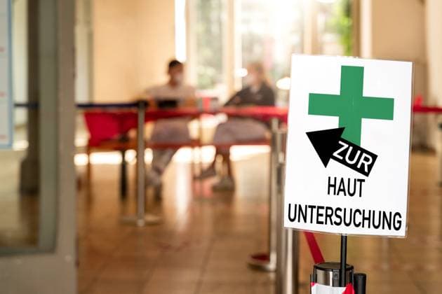 Zur HAUTUNTERSUCHUNG! Steht auf einem Schild, einer Arztpraxis, bzw. beim Hautarzt. Zu sehen sind medizinische Fachangestellte beim Facharzt. Symbolisiert Haut-Screenings, die Auffälligkeiten und Hautkrebs möglichst in einem frühen Stadium zu erkennen. FOTOMONTAGE