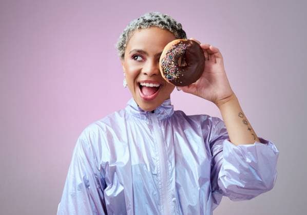 Creative Highlights Symbolfotos Fashion, donut and black woman excited and happy about dessert with futuristic vaporwave or holographic style clothing against purple background. Face of a happy female model looking cool and trendy. model released property released Copyright: xwww.peopleimages.comx