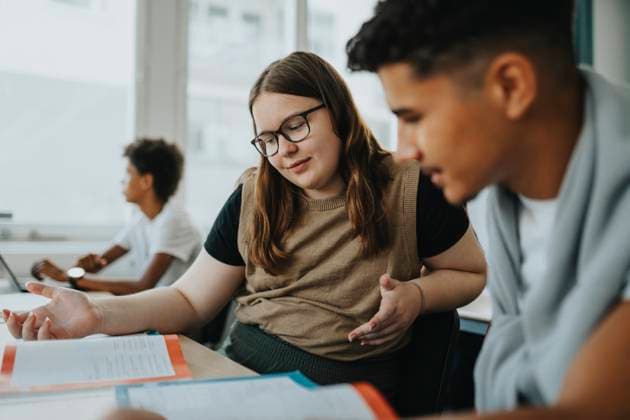 Teenage girl gesturing while discussing with male friend reading book at desk in classroom Sweden, Gothenburg, model released, property released Copyright: xMaskotx , MA113367 MA113367