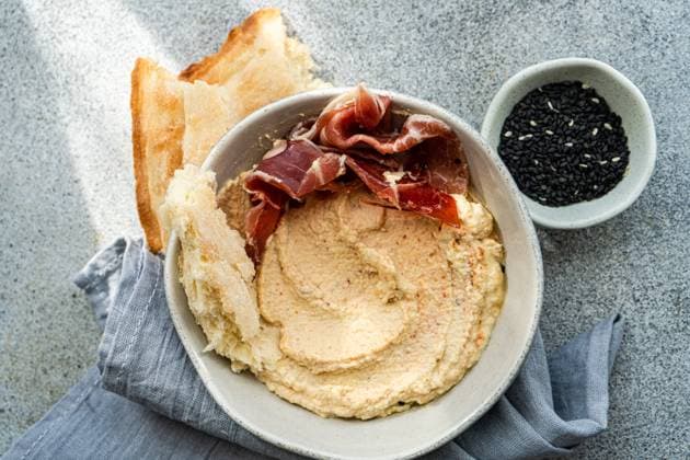 IMAGO Creative, Food, Tapas Top view of a ceramic bowl filled with hummus topped with slices of jamon, accompanied by toasted bread and a side dish of black sesame seeds, presented on a textured grey surface. Model Released AnnaBogush_ID24862_720224_007 Copyright: xAnnaxBogushx RECORD DATE NOT STATED