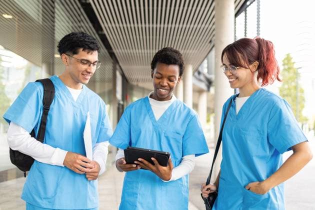 IMAGO Creative, Diversity, Pflege im Krankenhaus Diverse group of nursing students, two women and a man, engage in a lively discussion using a tablet outside a modern building, dressed in blue scrubs and carrying backpacks Model Released DavidMunoz_ID24882_720920_015 Copyright: xDavidxMunozx RECORD DATE NOT STATED