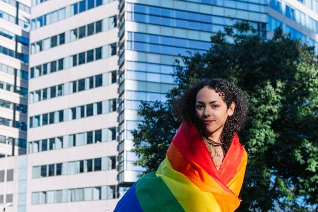 IMAGO Creative, Diversity, Teenager: LGBTQIA+ A joyful young trans woman with curly hair smiles while wrapped in a colorful rainbow flag, standing against a backdrop of modern buildings Model Released EmilioManzaneque_ID24620_713494_012 Copyright: xEmilioxManzanequex RECORD DATE NOT STATED