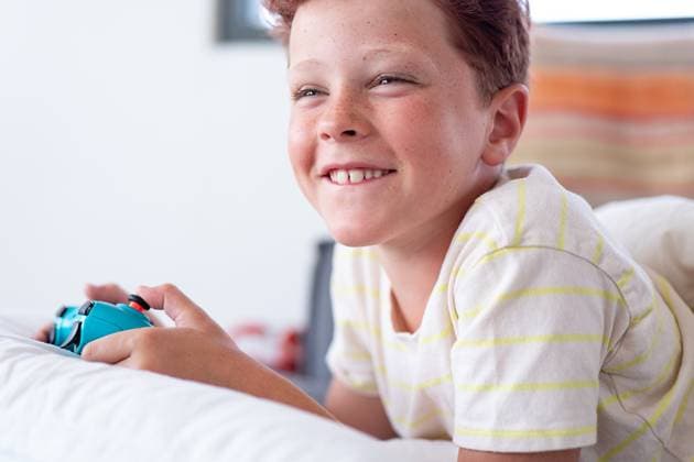 IMAGO Creative, Lifestyle, Gaming Young boy with red hair, visible freckles, and a big smile, playing video games on his handheld game console while lying on his stomach. He is wearing a light yellow striped t-shirt and looking slightly away from the camera. Model Released HodeiUnzueta_ID24741_716993_014 Copyright: xHodeixUnzuetax RECORD DATE NOT STATED