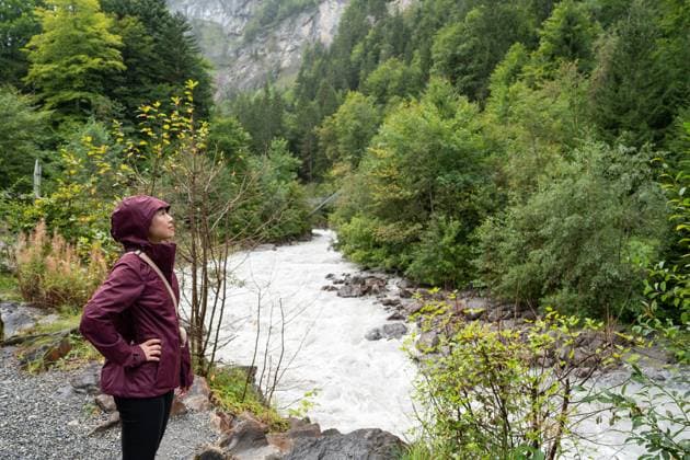 An asian woman in a burgundy raincoat stands beside a tumultuous river, surrounded by the lush, dense forests Naturpark Blausee, expressing a deep connection with the breathtaking Swiss nature Model Released JuanAlbertoRuiz_ID24556_711859_004 Copyright: xJuanxAlbertoxRuizx RECORD DATE NOT STATED