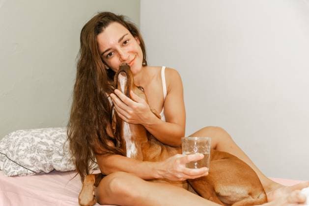 A young woman sits on a bed, lovingly holding her dog and a glass of water, sharing intimate moments of joy and care at home. Model Released NataliaPenado_ID24710_715927_024 Copyright: xNataliaxPenadox RECORD DATE NOT STATED